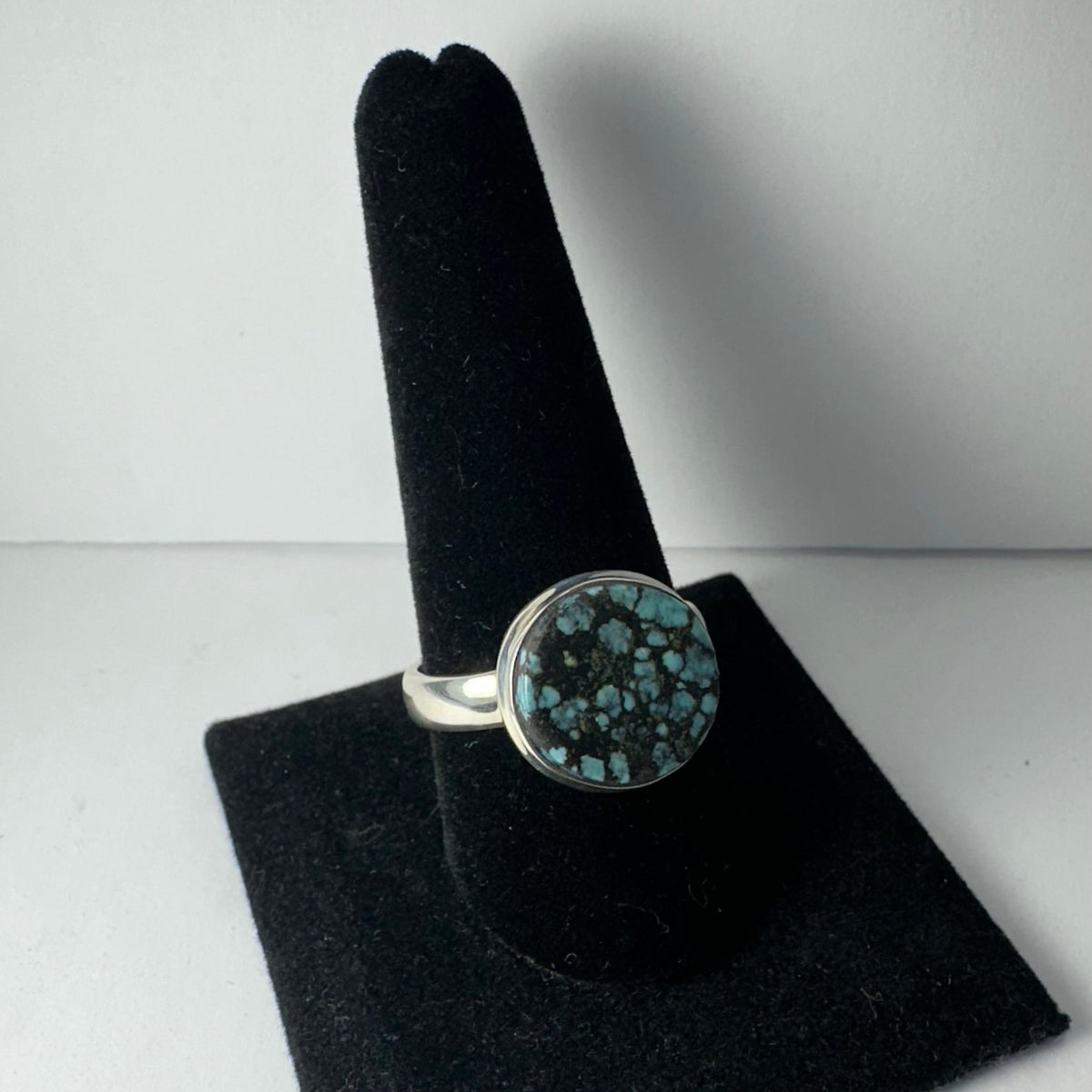 Silver ring with a round Tibetan turquoise stone displaying a mottled black and blue pattern, shown on a black velvet ring holder against a light gray background.