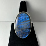 Oval labradorite ring with a silver bezel, displaying blue and gray iridescent streaks on a black velvet stand.