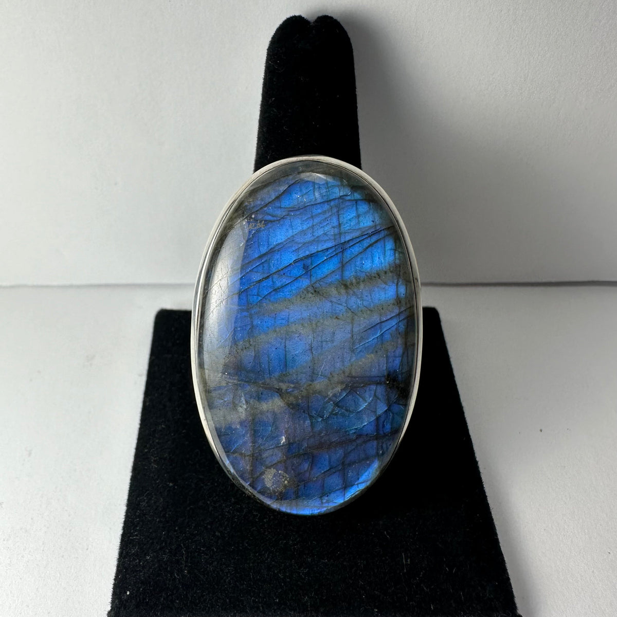 Oval labradorite ring with a silver bezel, displaying blue and gray iridescent streaks on a black velvet stand.