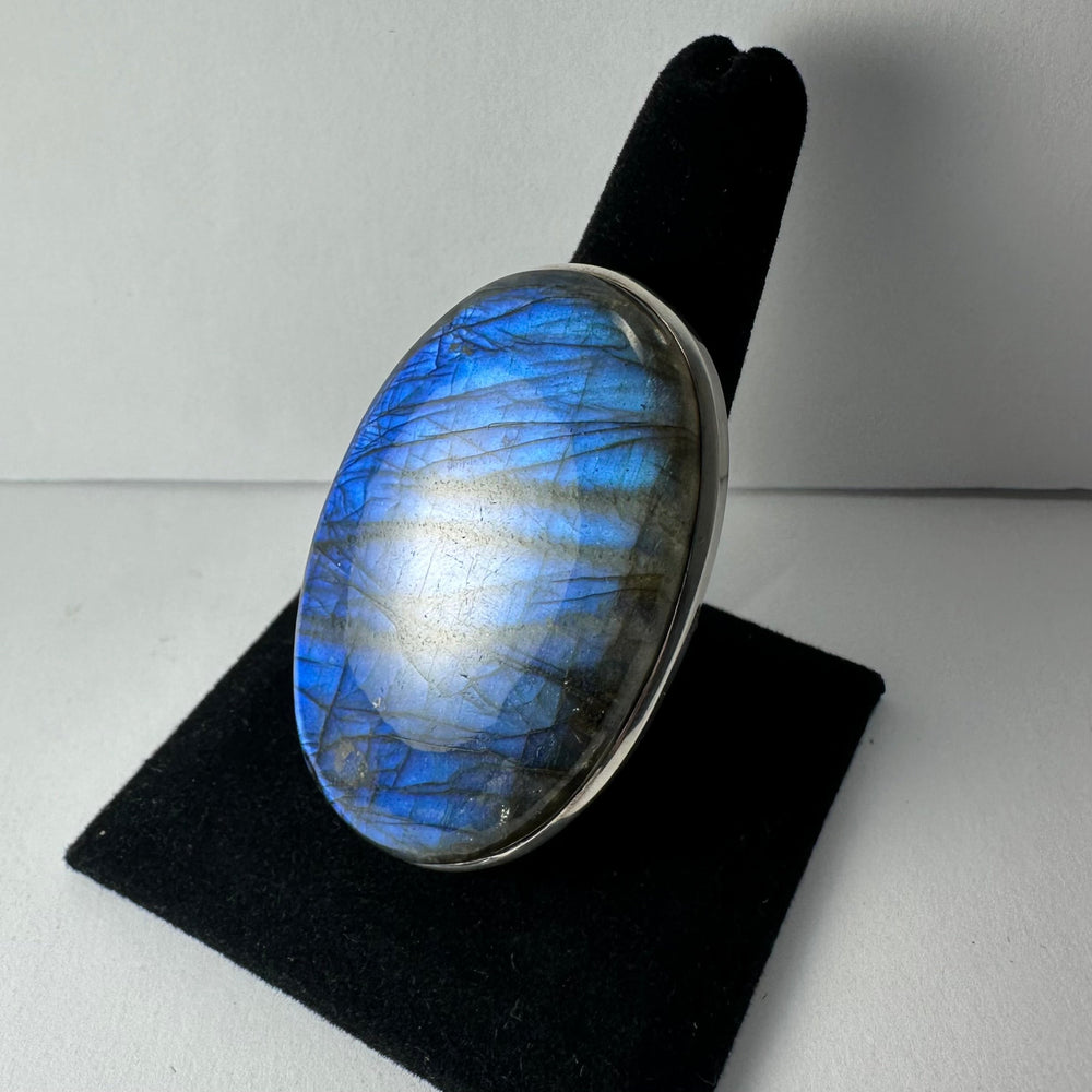 Oval labradorite gemstone ring with vibrant blue and gray iridescent flashes set in a simple silver bezel, displayed on a black velvet ring stand.