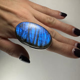 Large oval labradorite ring with vibrant blue and black striations worn on a hand with dark polished nails.
