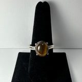 Silver ring with oval polished tiger eye gemstone set on a black velvet ring display stand, photographed against a light gray background.