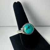 Silver ring with a round turquoise stone featuring black matrix patterns, displayed on a black velvet finger stand against a plain background.