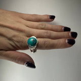Hand with dark purple nails wearing a silver ring featuring a round turquoise stone with natural black veining on the middle finger.