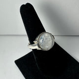Silver ring with a large, round, translucent moonstone set in a simple bezel, displayed on a black velvet stand against a plain white background.