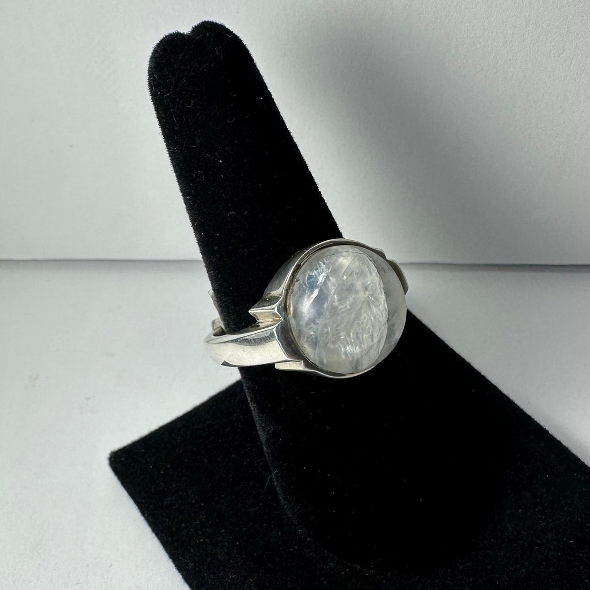Silver ring with a large, round, translucent moonstone set in a simple bezel, displayed on a black velvet stand against a plain white background.