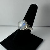 Round moonstone ring with silver band displayed on black velvet ring holder against a neutral gray background, showing subtle blue and white iridescence.