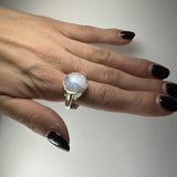 Hand with dark purple nail polish wearing a silver ring featuring a round, translucent moonstone with iridescent blue and white hues on the middle finger.