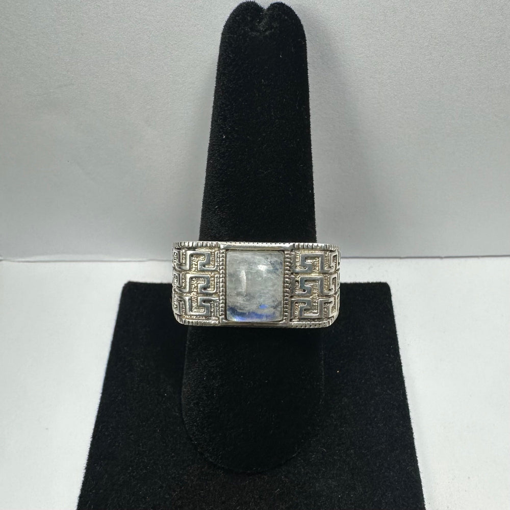 Silver ring with a rectangular moonstone center and intricate geometric patterns on the band, displayed on a black velvet ring stand.
