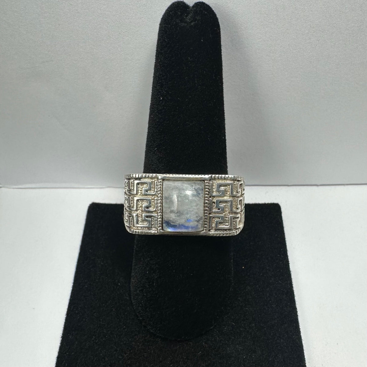 Silver ring with a rectangular moonstone center and intricate geometric patterns on the band, displayed on a black velvet ring stand.