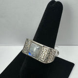 Silver ring with a square moonstone centered on a band featuring an intricate maze-like geometric pattern, displayed on a black velvet ring holder.