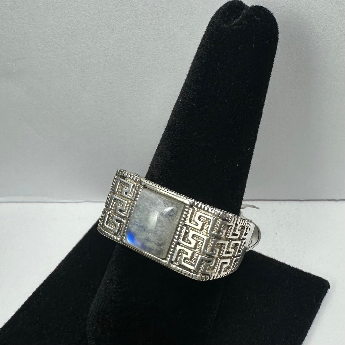 Silver ring with a square moonstone centered on a band featuring an intricate maze-like geometric pattern, displayed on a black velvet ring holder.