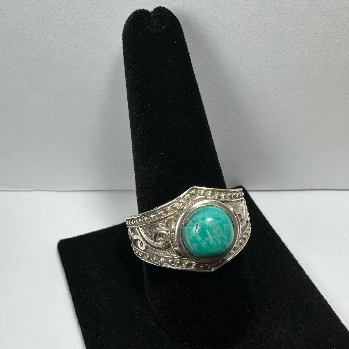 Silver ring with an ornate band featuring intricate scrollwork and a large round turquoise stone in the center, displayed on a black velvet ring holder.