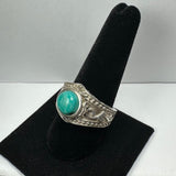 Silver ring with a round turquoise stone set in an ornately detailed band featuring embossed scroll patterns, displayed on a black velvet ring holder.