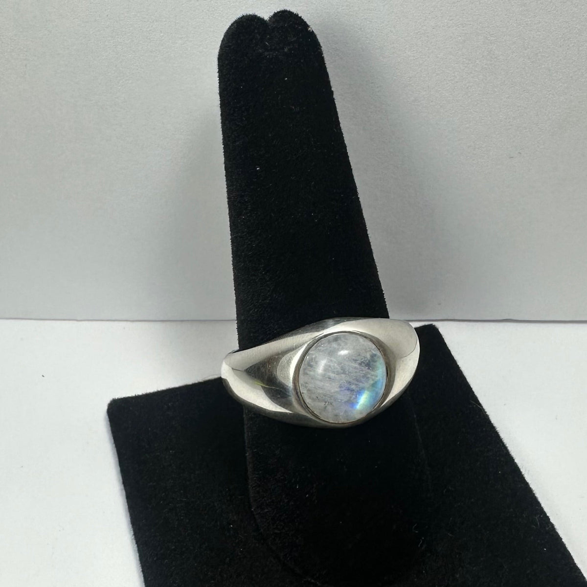 Silver ring with a round, translucent moonstone featuring blue iridescence, displayed on a black velvet ring holder against a plain gray background.