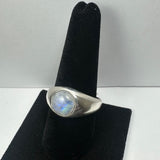 Silver ring with a rounded smooth moonstone that shows blue and iridescent flashes, mounted on a black velvet ring holder against a plain white background.
