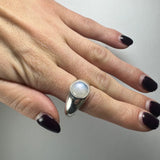 Hand wearing a silver ring with a round moonstone featuring a soft blue sheen, shown against a plain light background and dark purple nail polish.