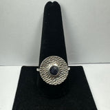 Silver ring with a hammered textured circular face featuring a small round dark blue iolite gemstone in the center, displayed on a black velvet stand.