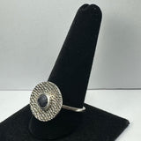 Silver ring with a round, textured disk setting featuring a dark blue oval iolite gemstone, displayed on a black velvet ring stand