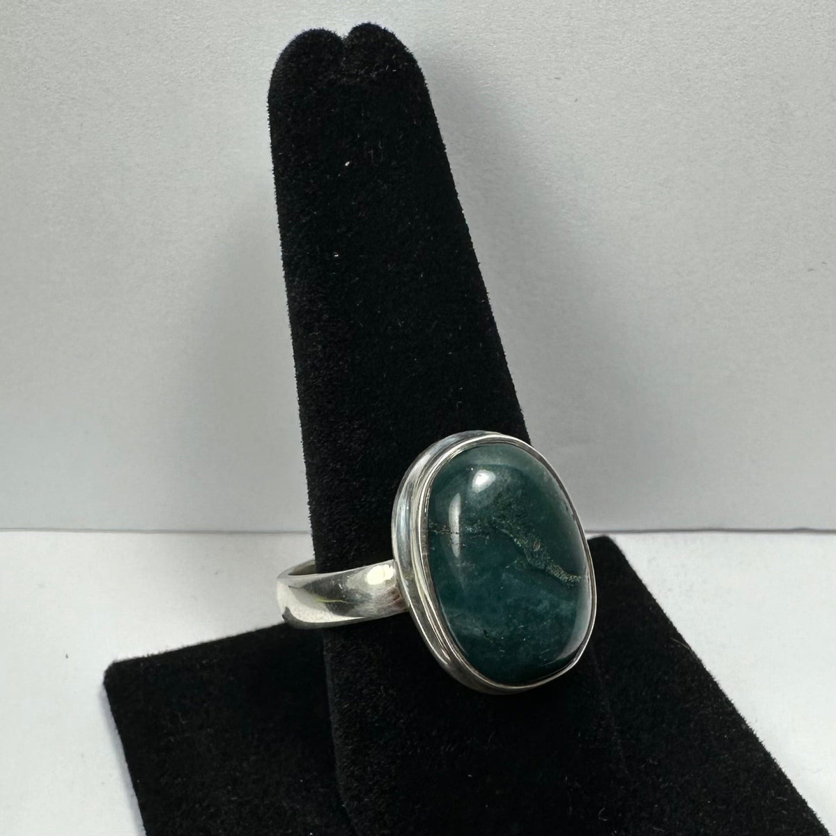 Silver ring featuring a polished oval green serpentine stone with subtle natural veining, displayed on a black velvet ring holder against a gray background.