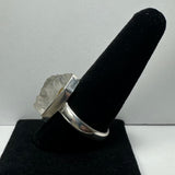 Silver ring with a large natural rough quartz crystal mounted on a wide band, displayed on a black velvet ring holder against a plain light background.