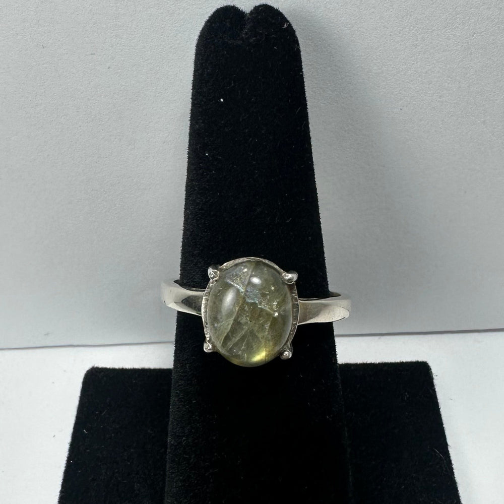 Silver ring with an oval labradorite stone exhibiting greenish-gray hues and natural inclusions, displayed vertically on a black velvet holder against a white background.
