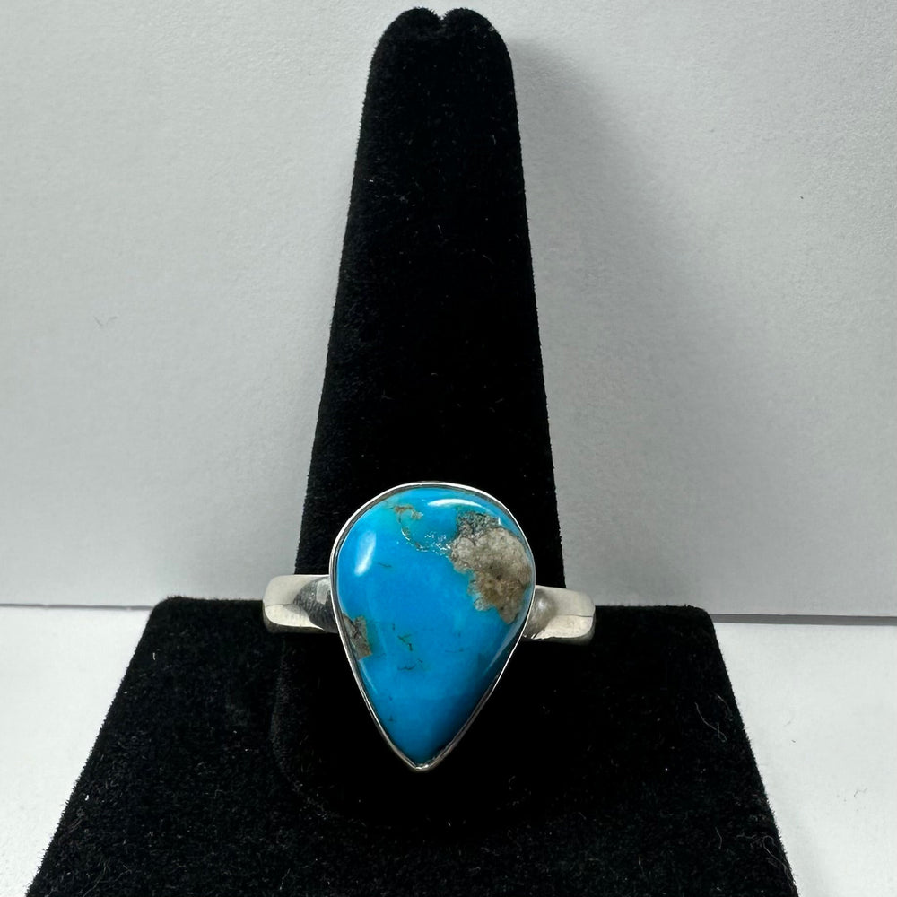 Silver ring with a teardrop-shaped vivid turquoise stone featuring natural brown matrix, displayed upright on a black velvet ring holder.
