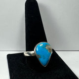Silver ring with a teardrop-shaped turquoise stone featuring natural brown matrix, displayed on a black velvet ring stand against a plain background.