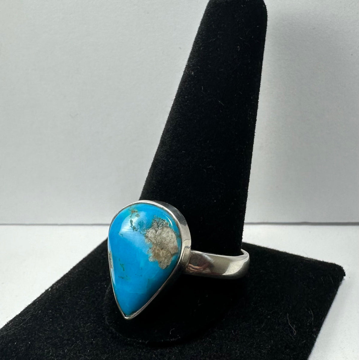Silver ring with a teardrop-shaped turquoise stone featuring natural brown matrix, displayed on a black velvet ring holder against a plain background.