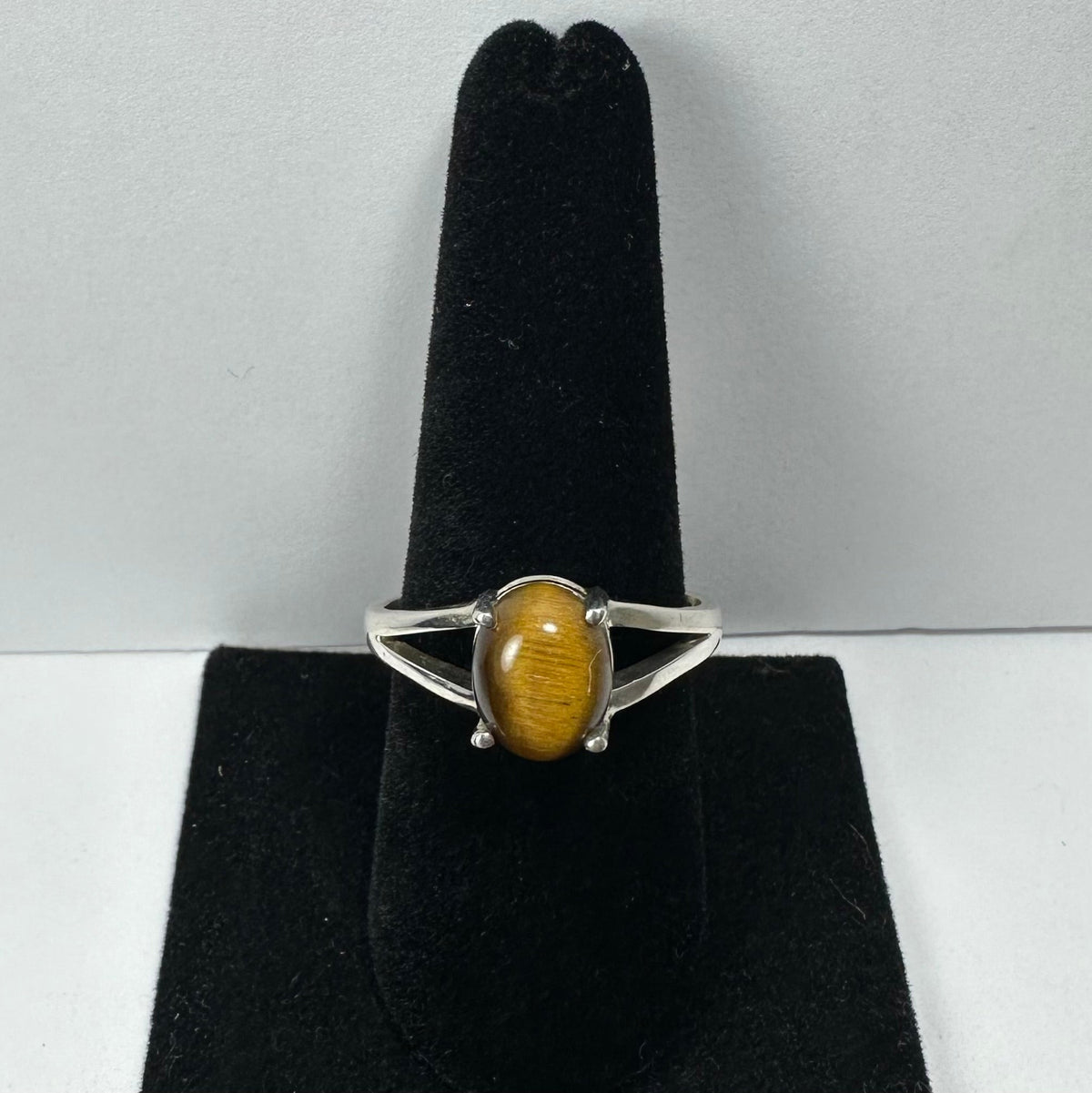 Silver ring with an oval polished tiger eye stone, displayed upright on a black velvet ring holder against a plain light background.