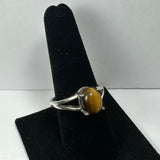 Silver ring with an oval tiger eye gemstone, featuring a split band design, displayed on a black velvet ring holder against a neutral background.