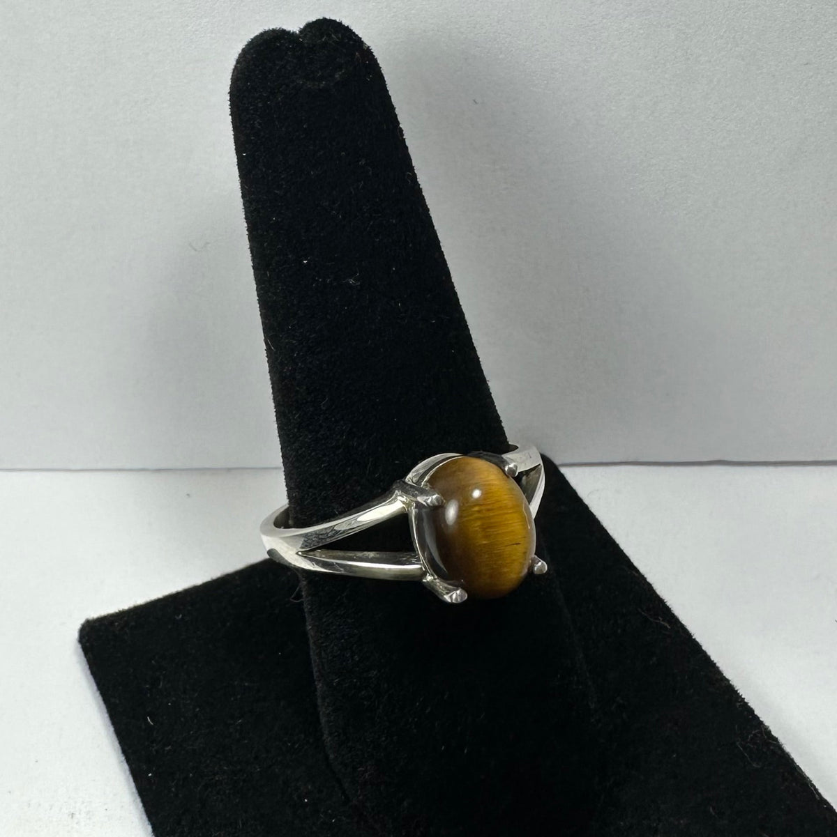 Silver ring with an oval tiger eye gemstone, featuring a split band design, displayed on a black velvet ring holder against a neutral background.
