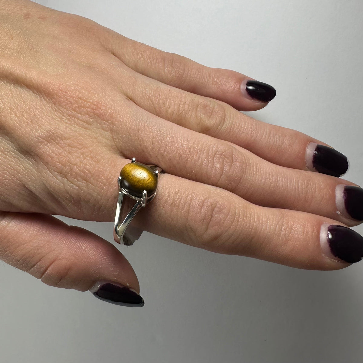Hand wearing a silver ring with a polished oval tiger eye stone, featuring a split shank band and dark purple nail polish on fingertips.