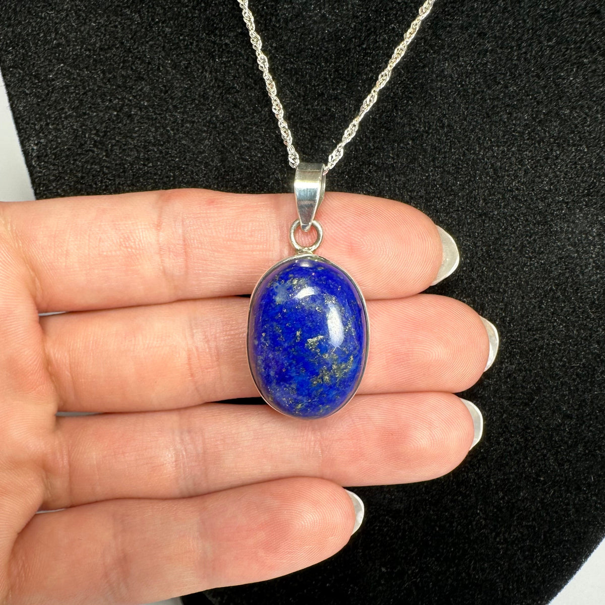 Oval lapis lazuli pendant with deep blue stone and gold flecks, set in silver and held by fingers against a black textured background.