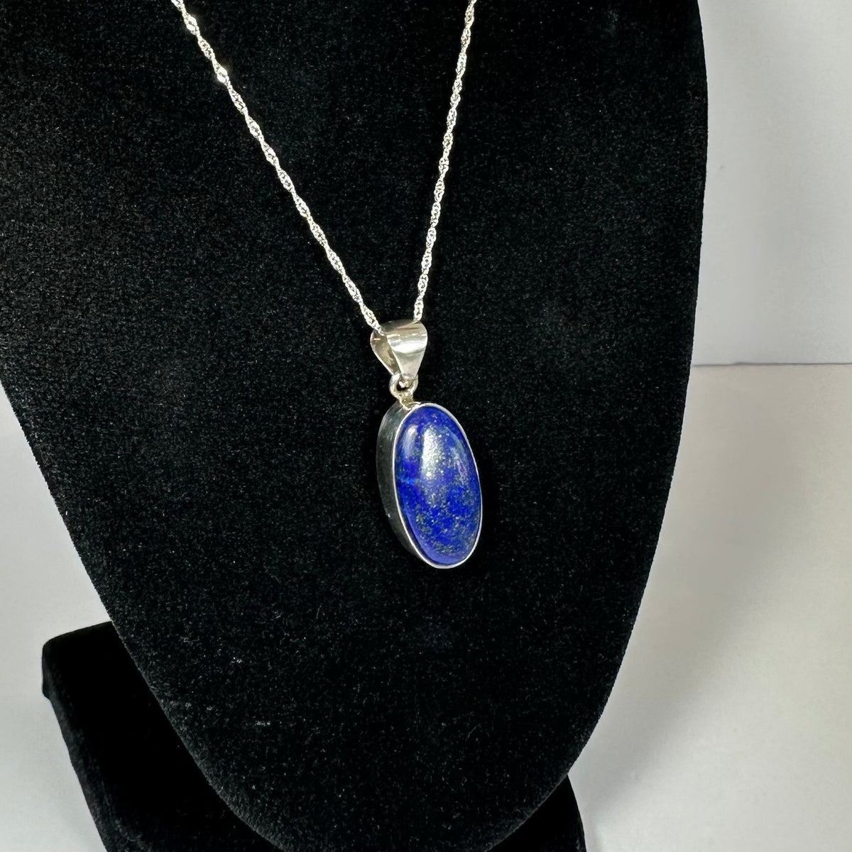 Oval lapis lazuli pendant with deep blue and gold flecks set in a silver bezel on a twisted silver chain against black velvet.