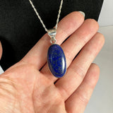 Oval-shaped deep blue lapis lazuli pendant with gold flecks set in a smooth silver frame, held in a hand against a black background.