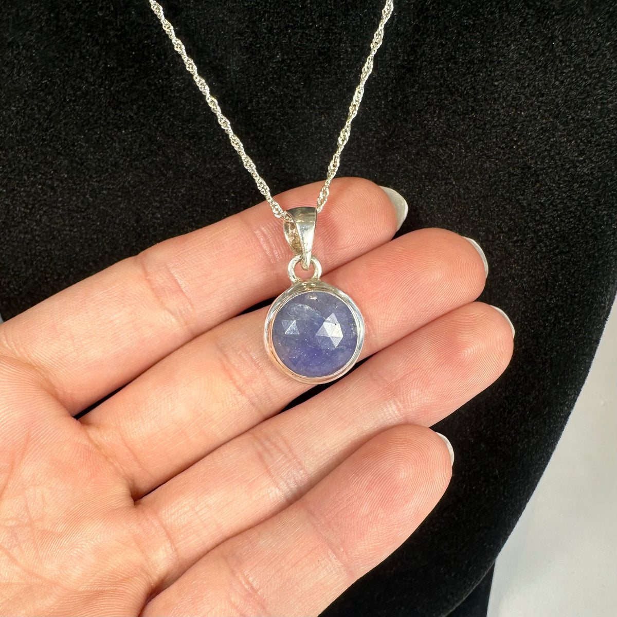 Round faceted tanzanite pendant set in a silver bezel on a delicate twisted silver chain, held against a black velvet background.