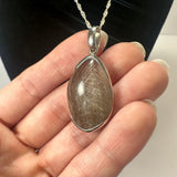 Teardrop-shaped pendant with silver rutile inclusions in a transparent stone, set in a silver bezel, held against fingers with a silver chain loop.