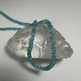 Blue apatite chip bead necklace loosely draped over a translucent, irregularly shaped crystal on a white background.