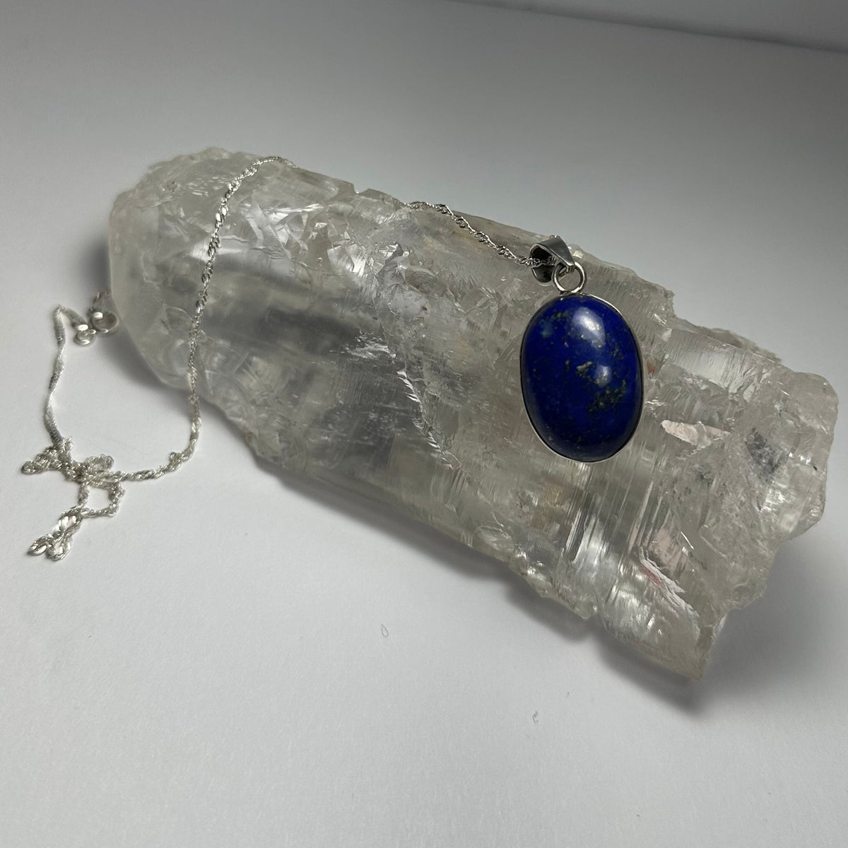 Oval lapis lazuli pendant with deep blue and gold flecks hanging on a delicate silver chain, displayed draped over a textured clear quartz crystal.