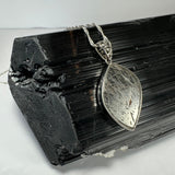 Marquise-shaped rutilated quartz pendant with silver filigree bail resting on a textured black tourmaline crystal cluster under soft lighting.