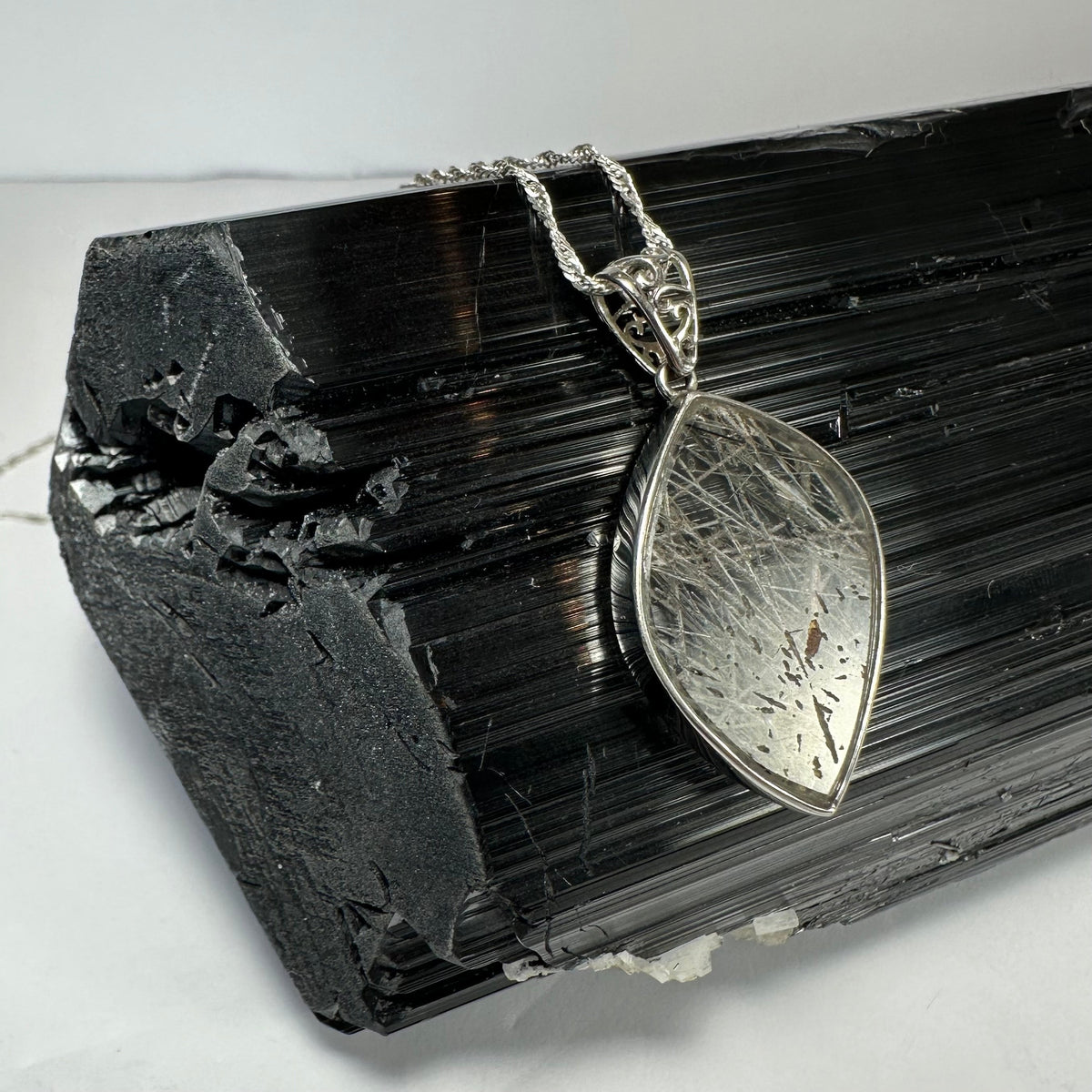 Marquise-shaped rutilated quartz pendant with silver filigree bail resting on a textured black tourmaline crystal cluster under soft lighting.