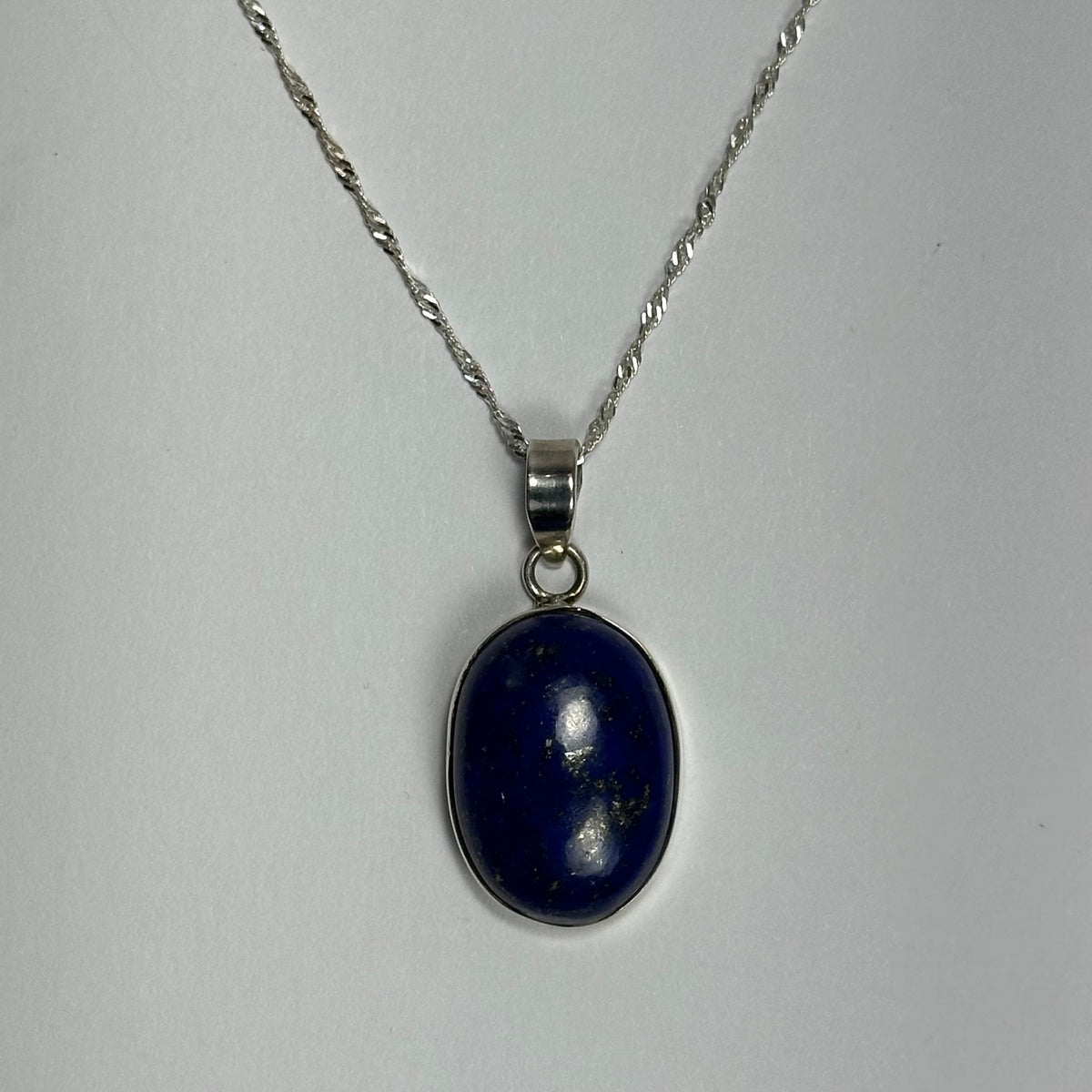 Oval lapis lazuli pendant with deep blue color and small gold flecks, set in silver and hanging from a twisted silver chain on a plain light background.
