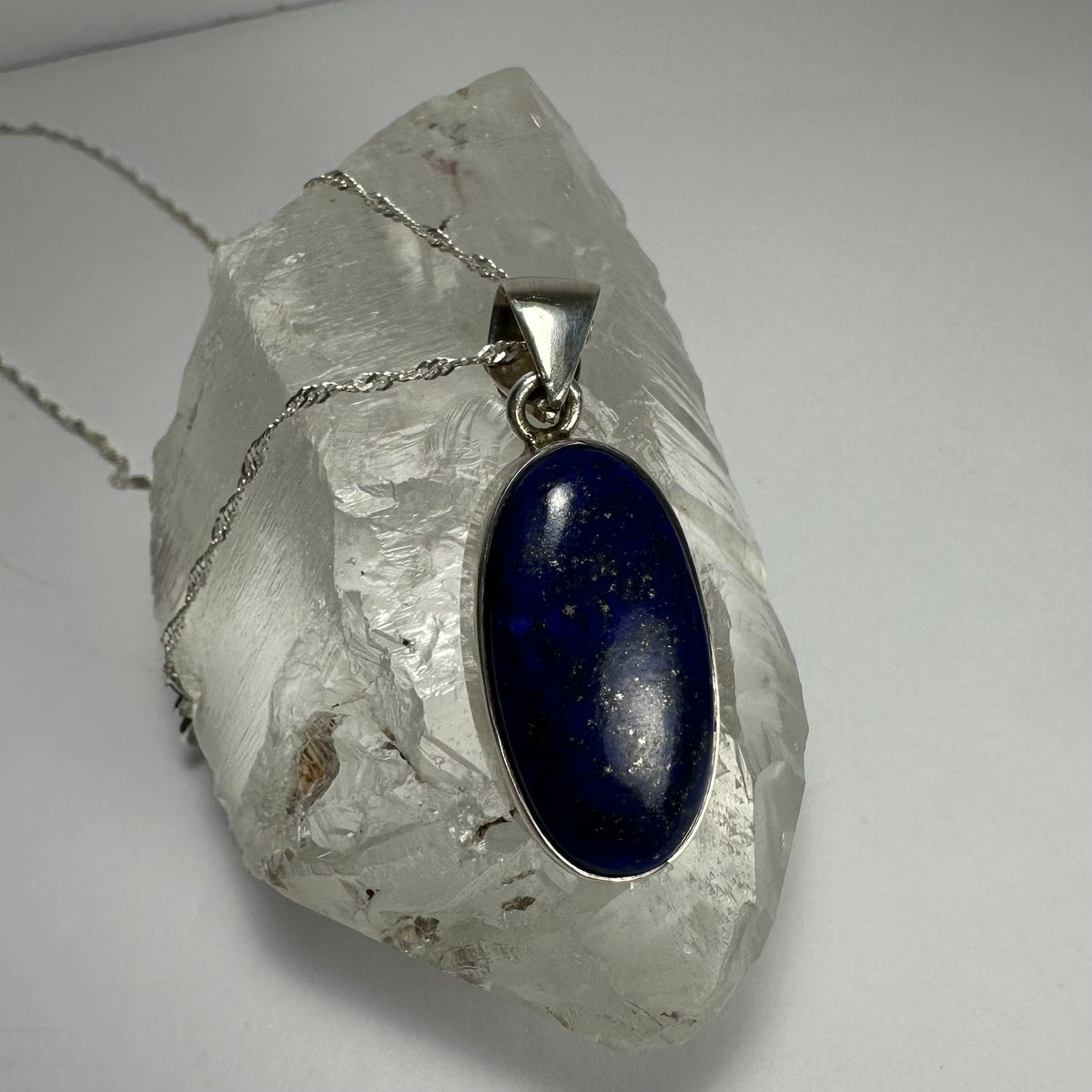 Oval deep blue lapis lazuli stone pendant with subtle gold flecks set in silver, resting on a large raw clear quartz crystal against a white background.