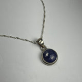 Silver chain necklace with a round faceted deep blue tanzanite pendant set in a smooth silver bezel, displayed on a plain light surface.