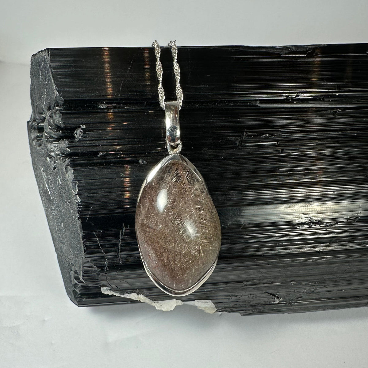 Oval silver pendant with translucent stone featuring fine silver rutile inclusions, displayed against a textured black mineral background, hanging from a twisted silver chain.