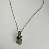Silver chain necklace with a small raw green tourmaline crystal pendant set in a simple silver bezel, displayed against a plain white background.