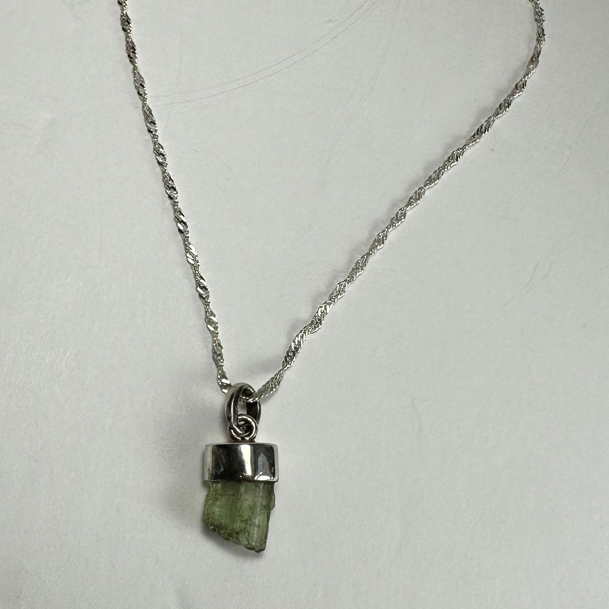 Silver chain necklace with a small raw green tourmaline crystal pendant set in a simple silver bezel, displayed against a plain white background.