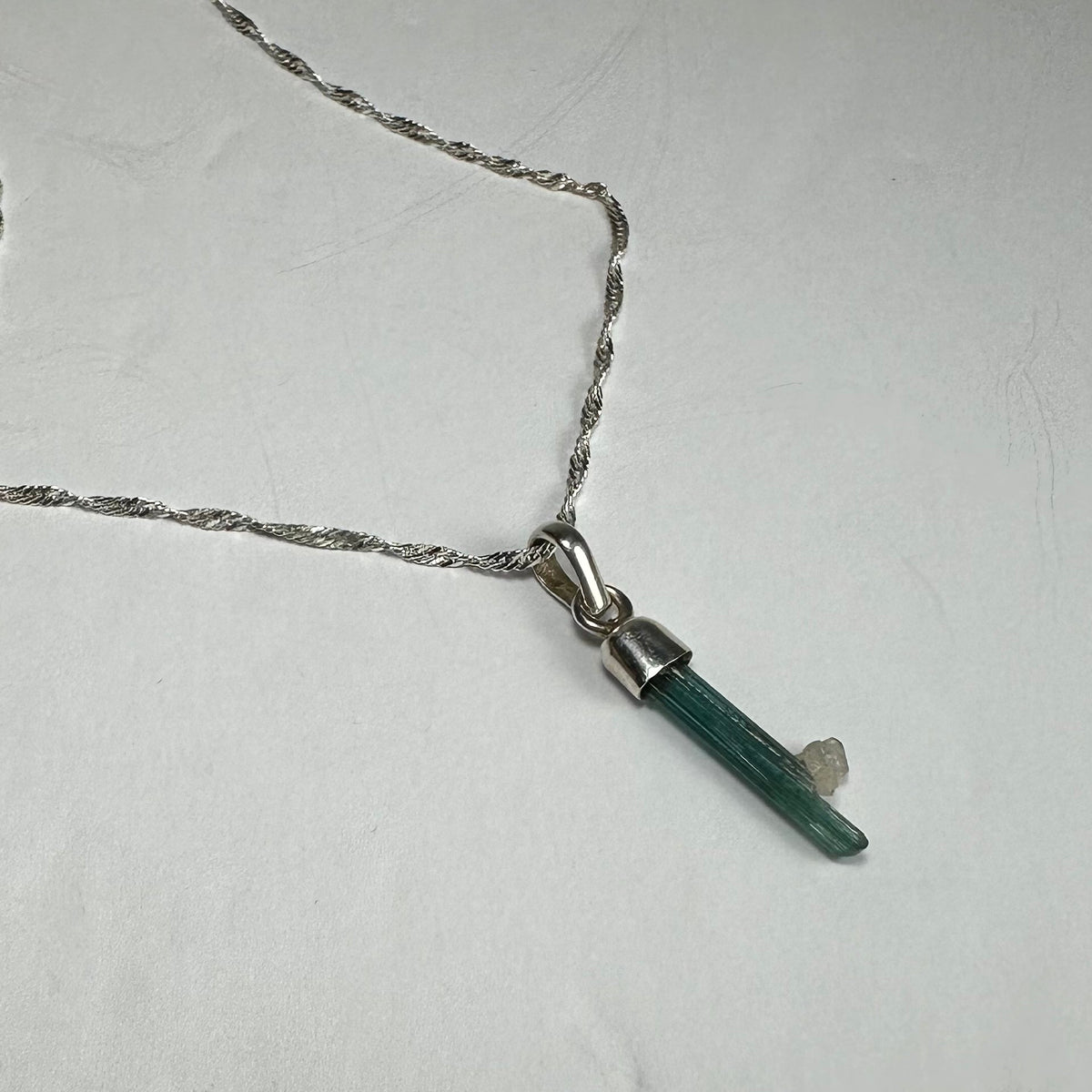 Silver chain necklace with a pendant featuring a slender, green tourmaline crystal with a small, rough white crystal attached.