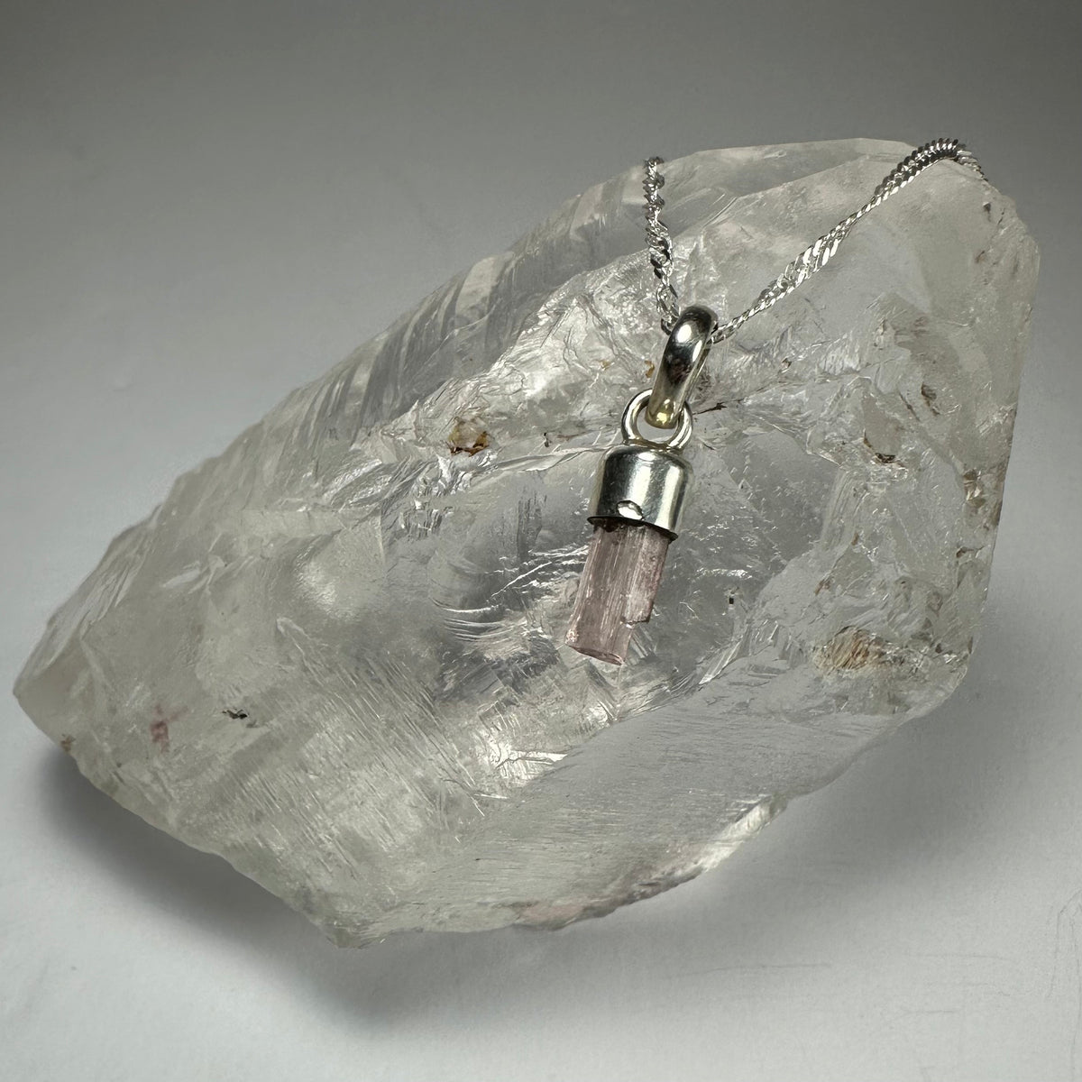 Small pale pink tourmaline crystal pendant with silver cap and chain, resting on a large clear quartz crystal with textured surface.